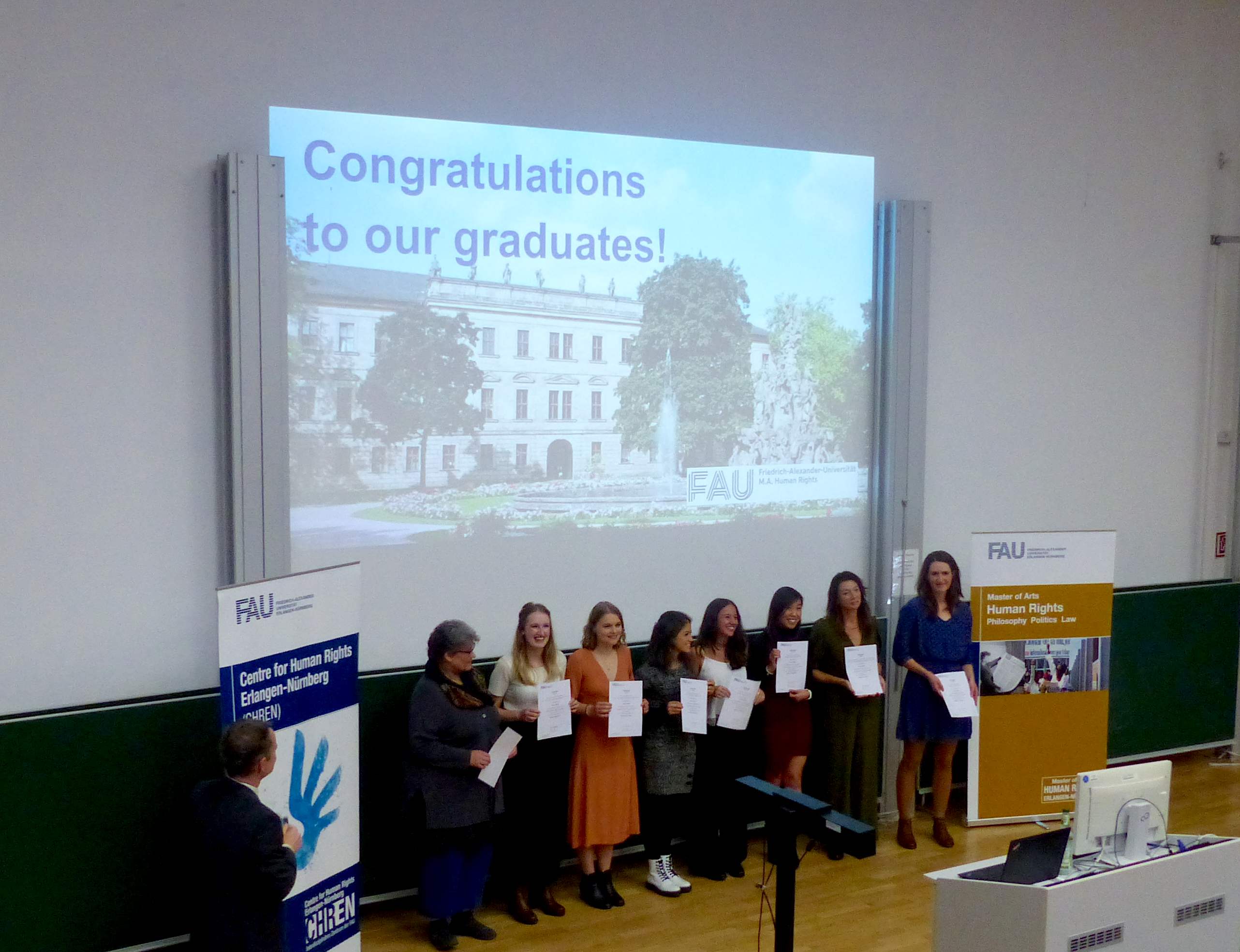 Picture of graduates at their graduation ceremony in front of a PowerPoint "Congratulations to our graduates!" holding their diplomas.
