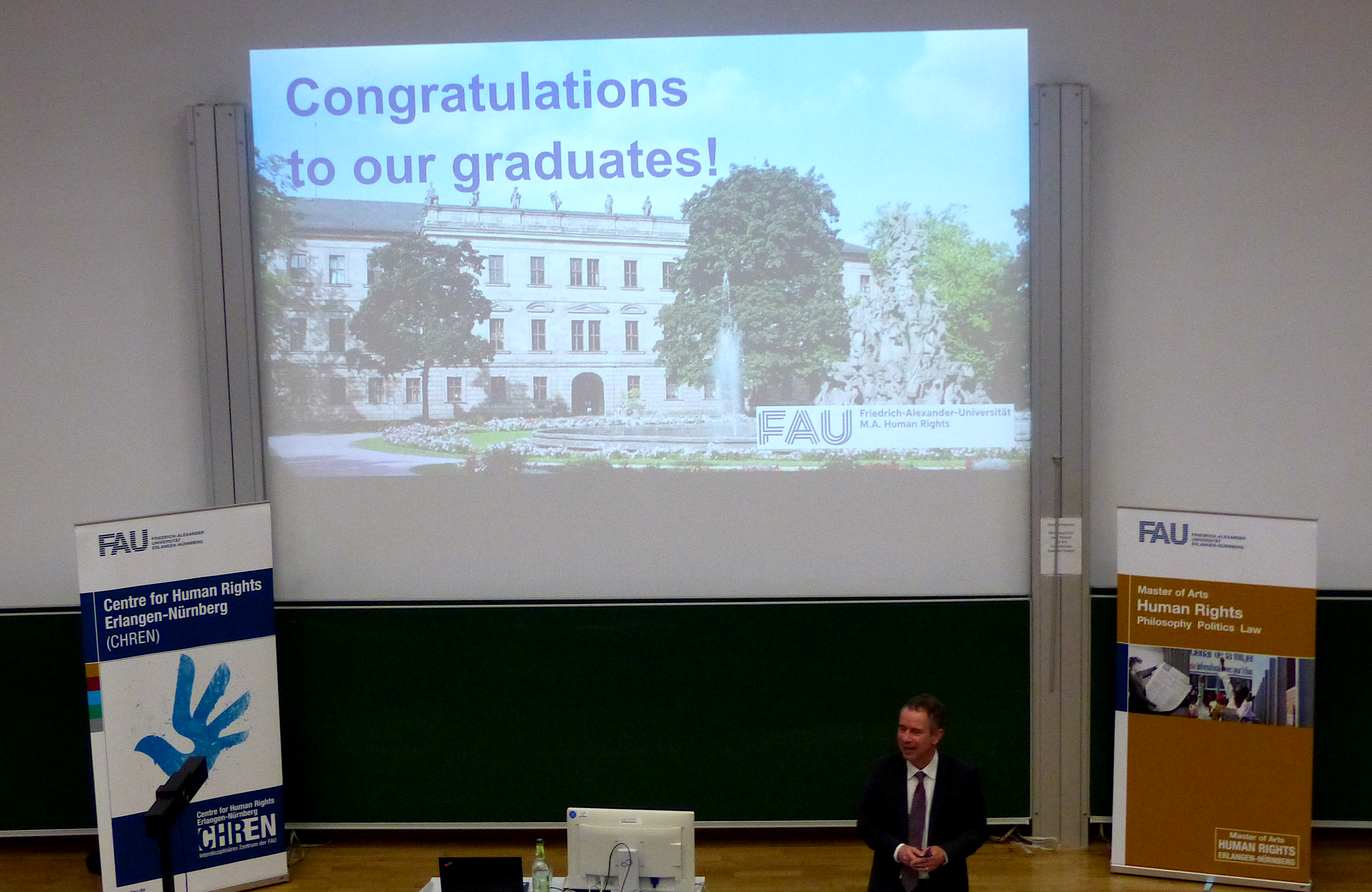 Picture of Prof. Krajewski at the graduation ceremony in front of a powerpoint "Congratulations to our graduates!"