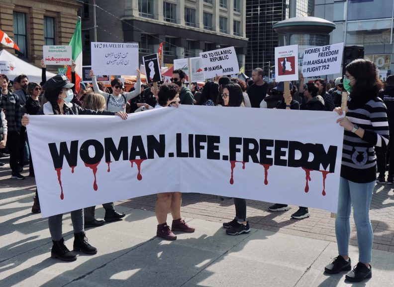 Picture of two women holding a white sign with big black letters from which blood is dropping down; Woman, Life, Freedom at a protest.